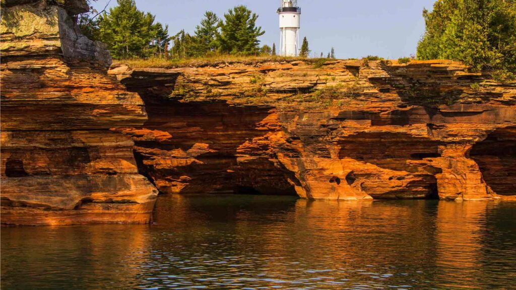 Striking red sandstone cliffs and a white lighthouse above the shoreline in Apostle Islands, WI — a hidden US destination known for sea caves and scenic kayaking.