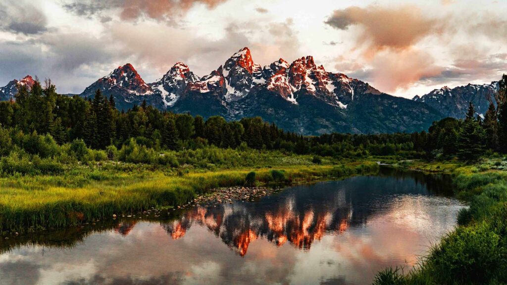 Majestic snow-capped peaks of the Grand Teton range glowing at sunset, reflected in a calm river surrounded by lush greenery—an unforgettable hidden US destination in Wyoming.