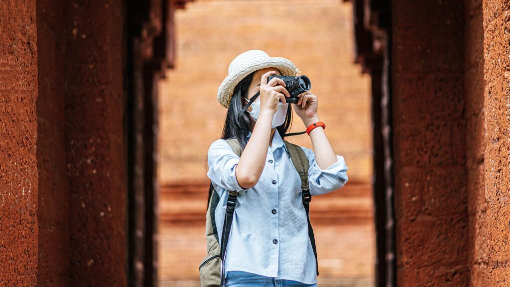 Solo traveler wearing a hat and mask, taking a photo at a historical site during a cultural solo travel adventure.