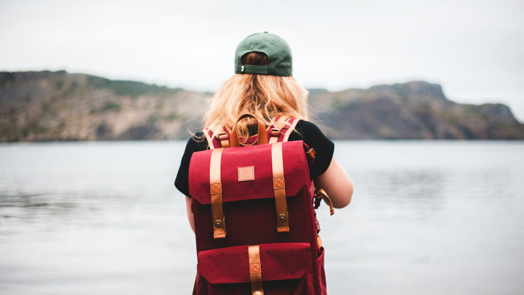 Woman with a red backpack facing a calm lake and mountains, symbolizing the freedom and adventure of solo travel.