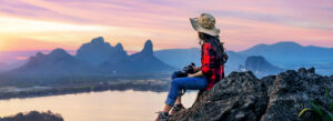 Woman enjoying a solo travel experience while sitting on a rocky viewpoint at sunset, overlooking mountains and a river.