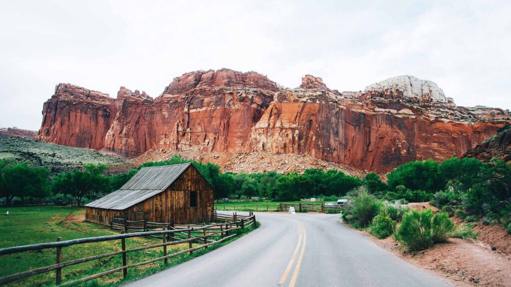 View of Scenic Byway 12 in Utah with a rustic barn, lush greenery, and towering red rock cliffs, highlighting one of the most breathtaking road trips in USA.