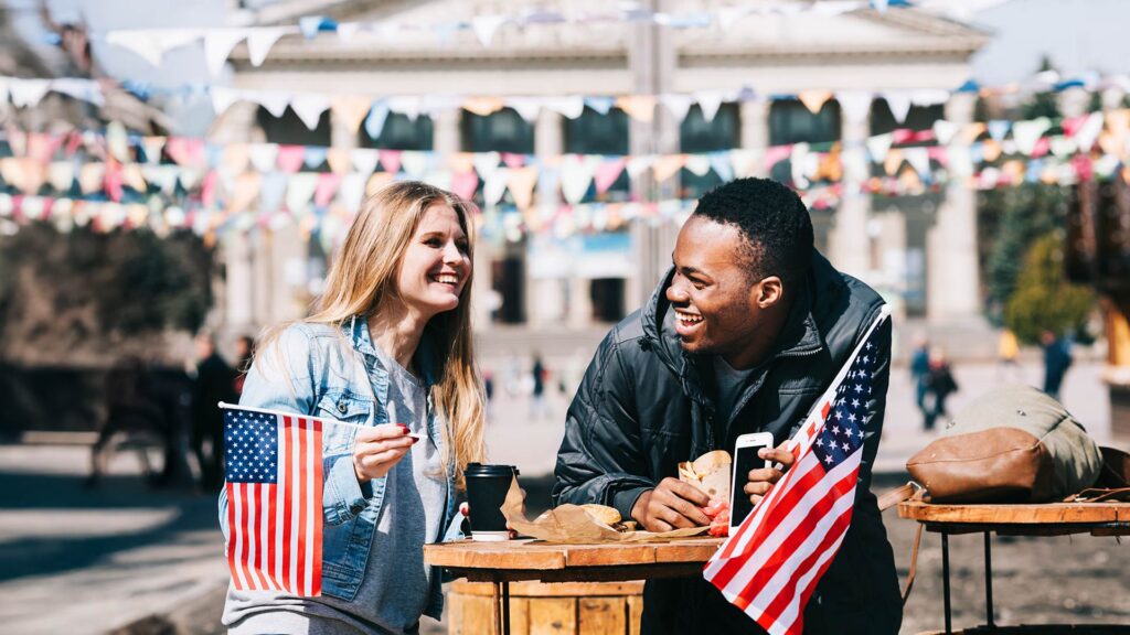 Happy young adults enjoying food and waving small American flags during a festive outdoor event for the 4th of July.