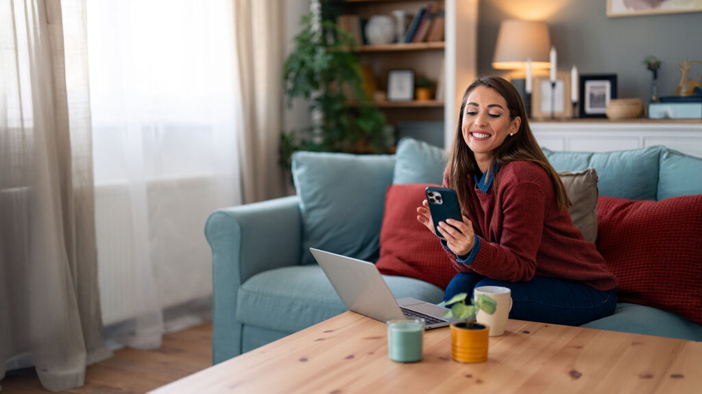 Smiling woman checking her phone and laptop at home, feeling confident after avoiding common booking mistakes