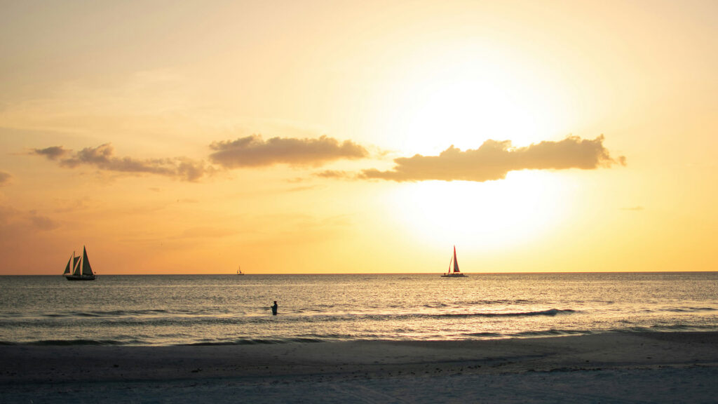 Golden sunset over Clearwater Beach, Florida with sailboats drifting across the calm sea and a person wading in the water—an inviting scene for affordable beach vacations.