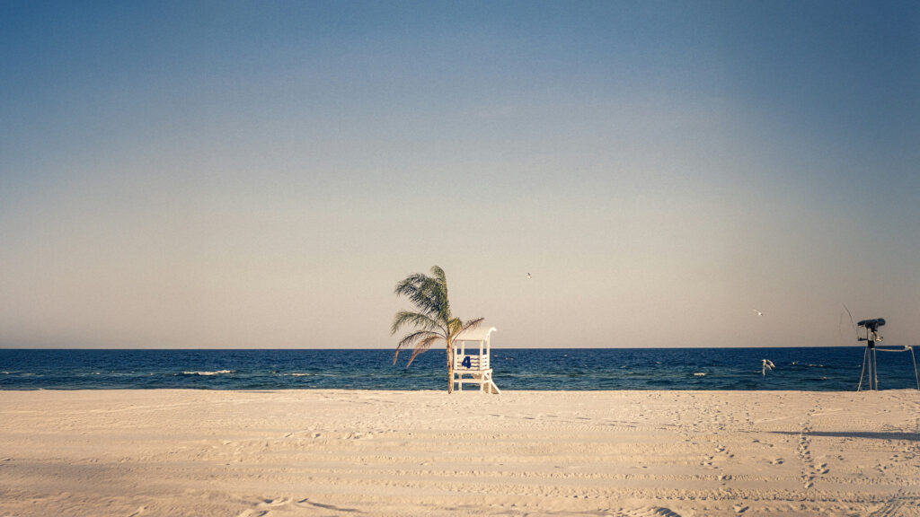 Scenic view of a quiet beach in Gulf Shores, Alabama with white sand, a lifeguard chair, and a palm tree near the shoreline under a clear sky — a perfect spot for affordable beach vacations.