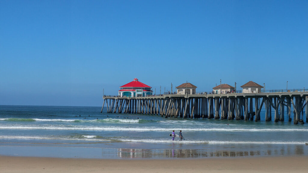 View of Huntington Beach Pier in California with a bright red-roofed building, surfers in the water, and beachgoers enjoying the shore—an ideal spot for affordable beach vacations on the West Coast.