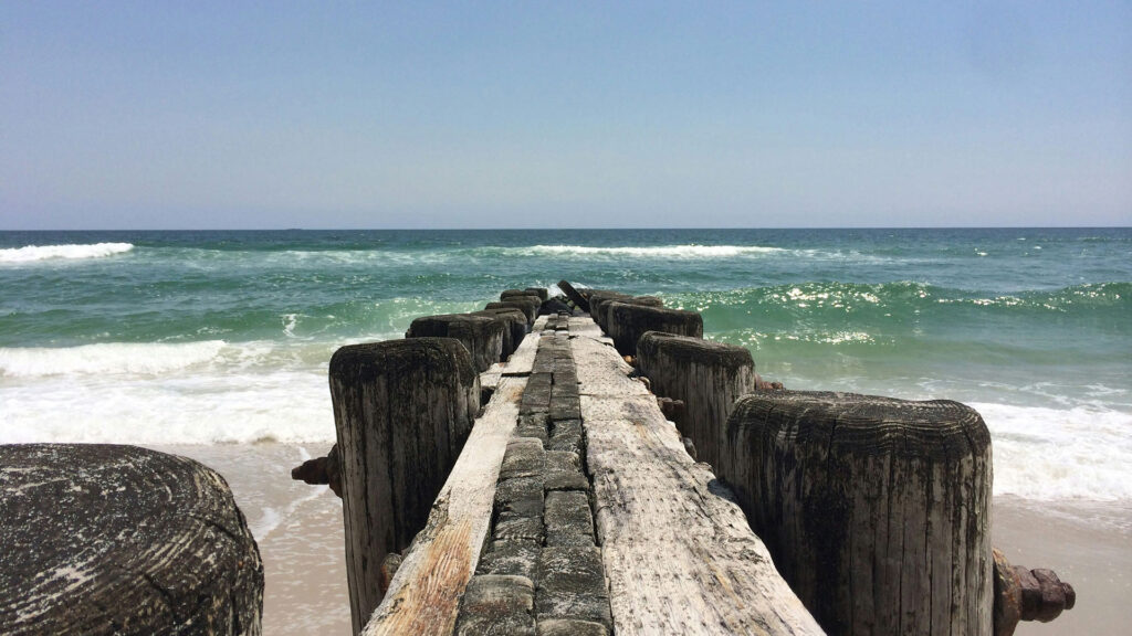 A weathered wooden boardwalk extends out into the ocean with waves crashing on a sandy beach under a clear blue sky—an inviting scene from the Outer Banks, North Carolina, perfect for affordable beach vacations.