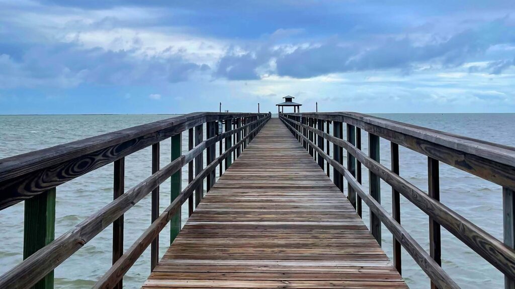 A long wooden pier stretches over calm ocean waters under a cloudy sky at Rockport Beach, Texas—an inviting and peaceful spot ideal for affordable beach vacations on the Gulf Coast.