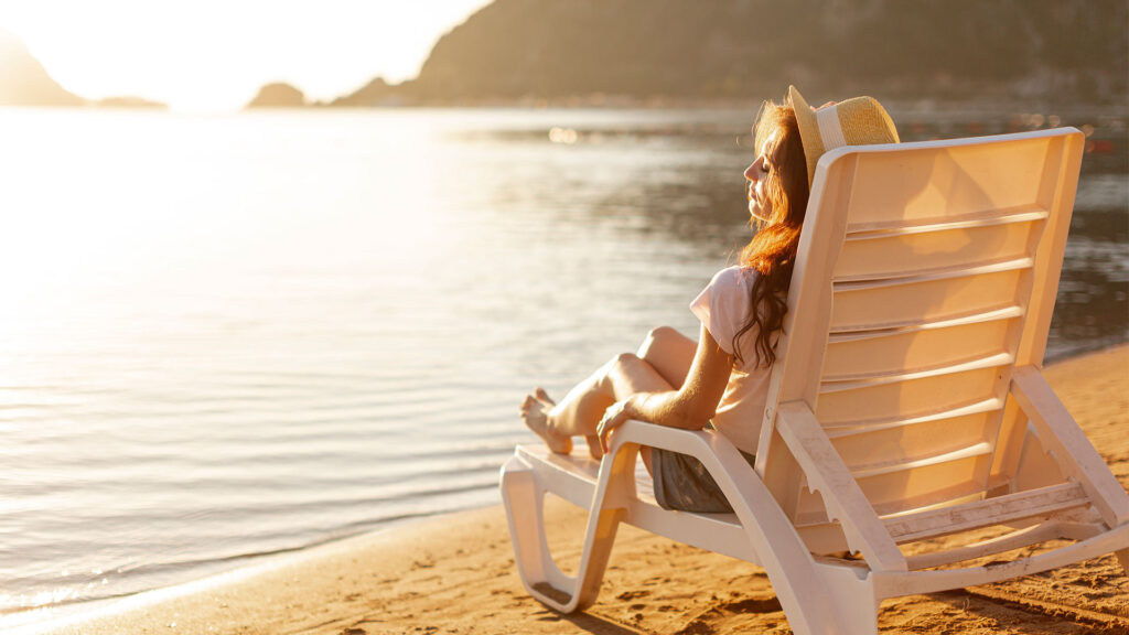 A woman relaxes on a lounge chair by the shoreline during sunset, enjoying a peaceful moment on the sand—perfect representation of affordable beach vacations that offer tranquility without the high price tag.