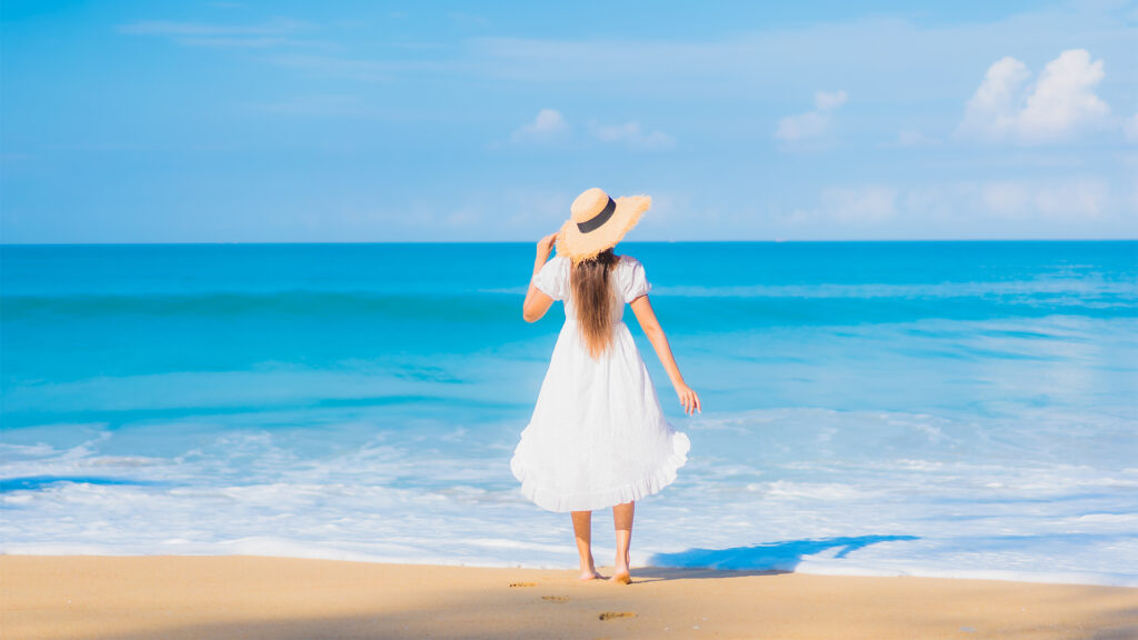 A woman in a white dress and straw hat, standing barefoot on the beach, facing the ocean with clear blue skies and gentle waves.
