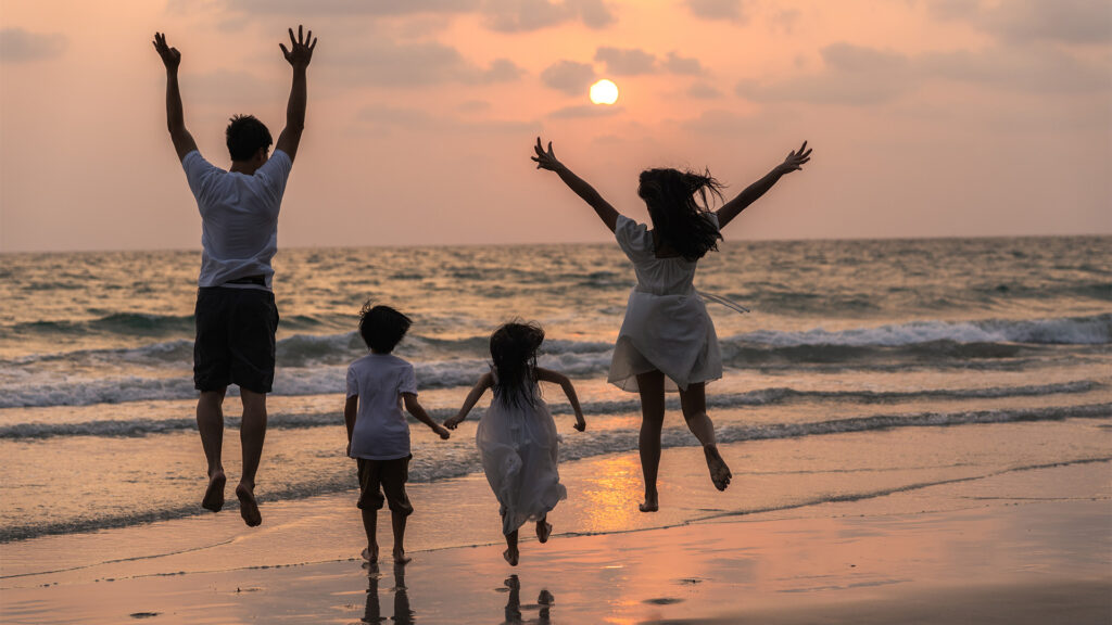 A family of four jumping joyfully on the beach during sunset, with the ocean and waves in the background.