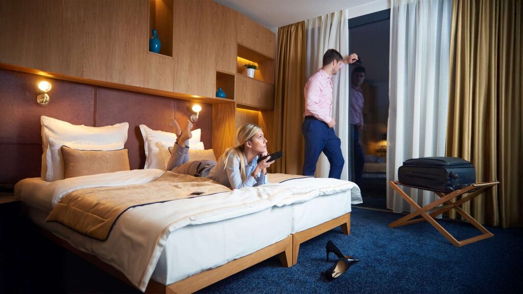 Business couple in a stylish yet cheap hotel room; woman watching TV on bed while man looks out the window.