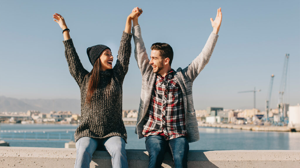 A couple raises their arms in excitement at a scenic harbor, enjoying the freedom and joy of their vacation.