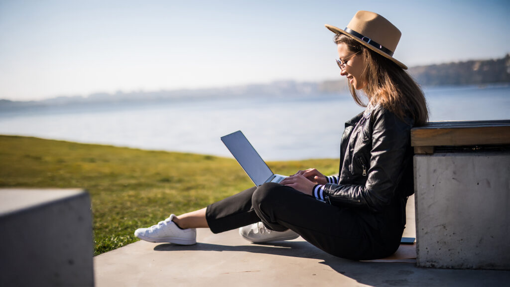 Woman working on a laptop outdoors, sitting on a concrete bench by a body of water, enjoying the scenic view.