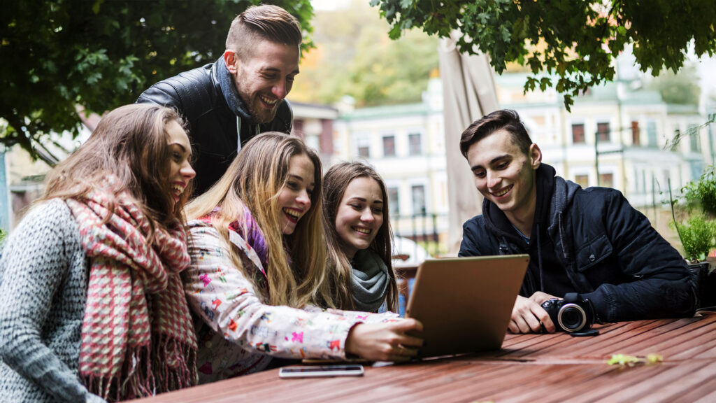 Group of five friends smiling and looking at a laptop together while sitting outdoors at a wooden table.