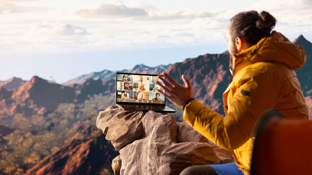 Person in a yellow jacket participating in a video call while sitting on a rock with a scenic mountainous background.