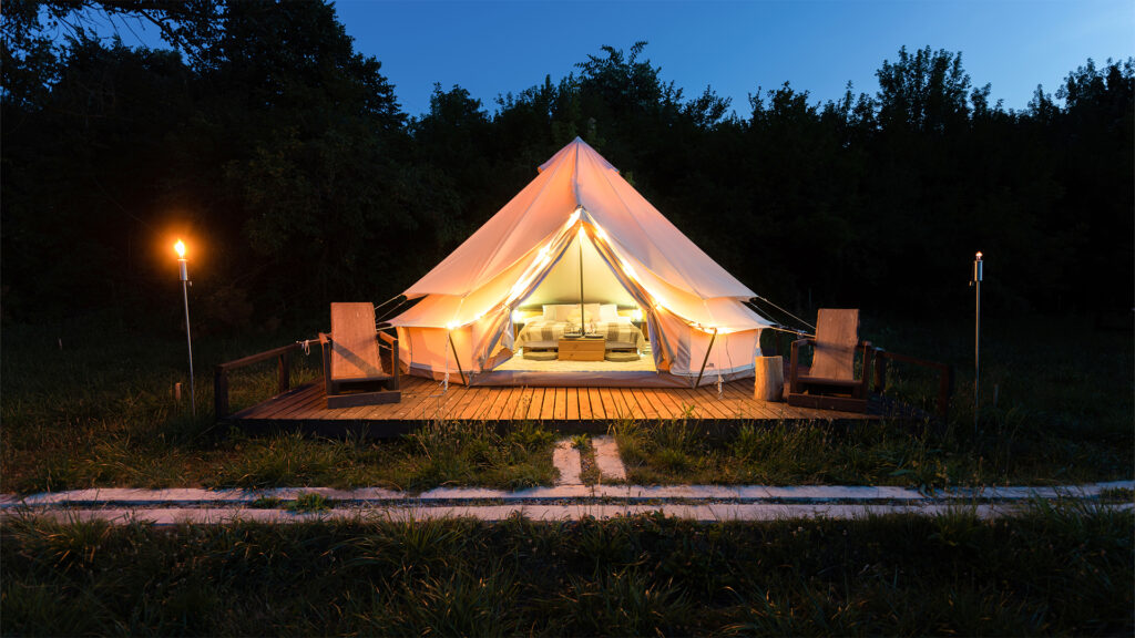 A luxurious glamping tent with a cozy bed inside, softly illuminated at dusk. The tent is set on a wooden deck surrounded by a grassy field, with torches lighting the path leading to it.