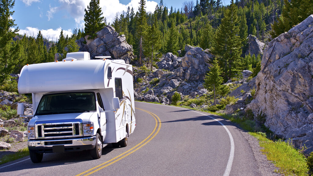 A white RV driving along a winding road through a lush forest, with towering rocky cliffs on the side under a bright blue sky.