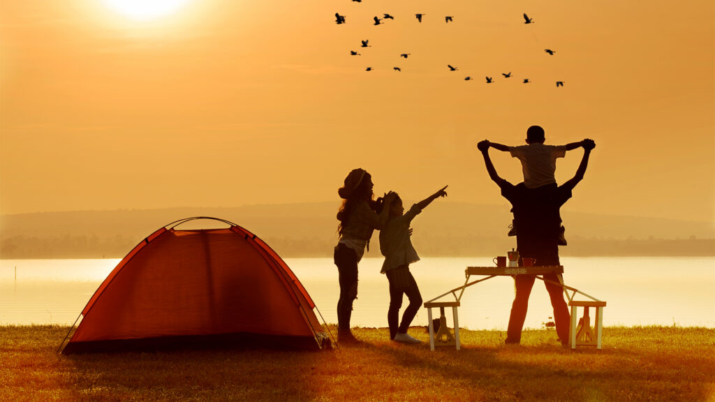 A family enjoying a sunset by a lake, with a red tent in the foreground. The family is playing together, with birds flying across the orange sky.