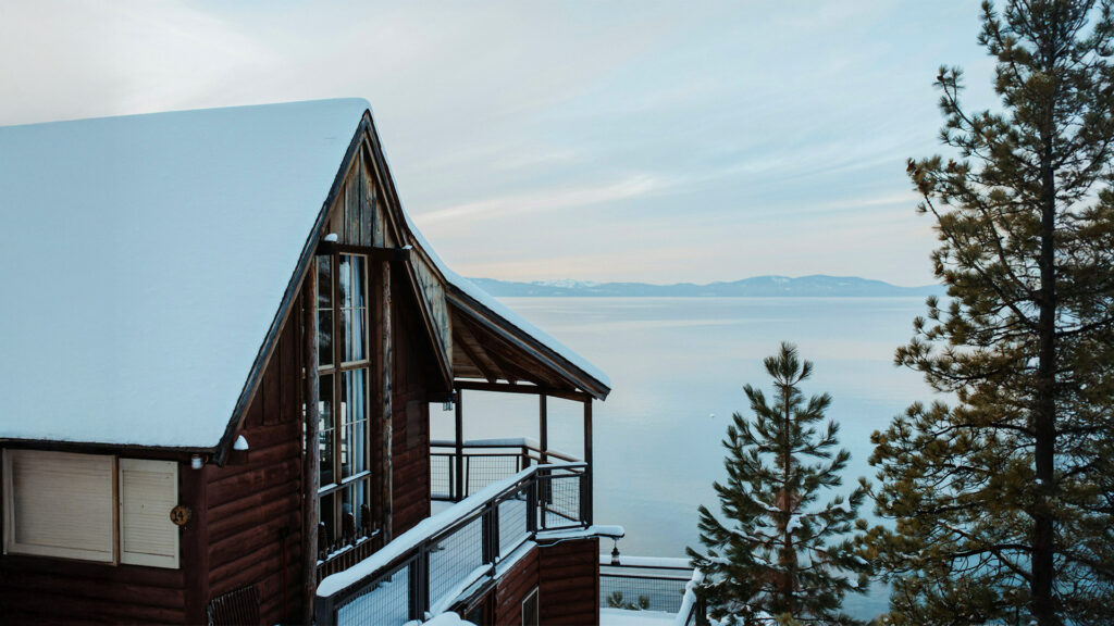 A cozy wooden cabin covered in snow, overlooking Lake Tahoe. The surrounding pine trees add to the winter charm, and the tranquil lake reflects the snowy landscape and distant mountains.