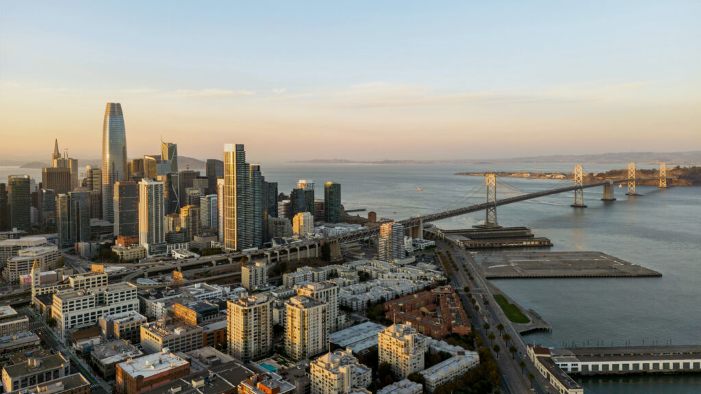 A stunning aerial view of the San Francisco skyline at sunset, showcasing towering modern skyscrapers, including the Salesforce Tower, and the iconic Bay Bridge stretching over the water. The city's streets and waterfront buildings are bathed in golden light as the sun sets in the distance.
