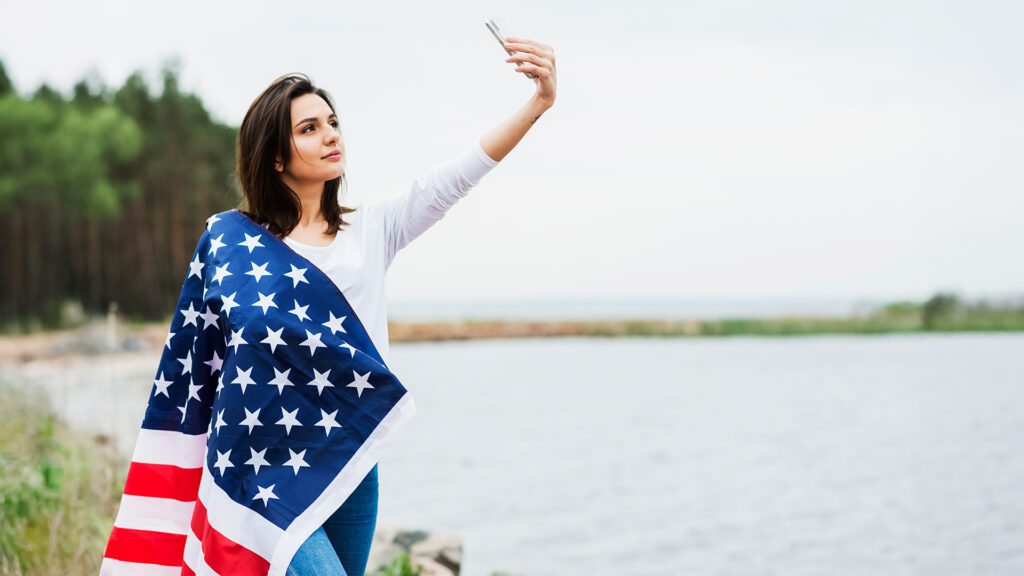 Woman wrapped in American flag taking selfie by the water on a travel journey.