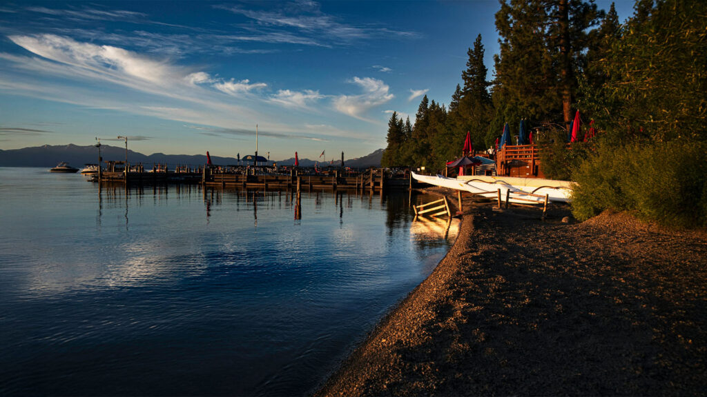 A peaceful view of the Lake Tahoe dock at sunset, with calm water reflecting the blue sky and mountains in the background. A row of boats is visible near the shore, with trees lining the horizon.