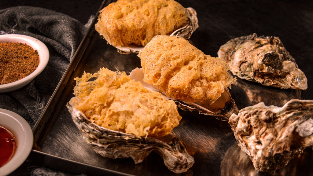 A serving of fried Rocky Mountain oysters with a golden, crispy crust served on a metal tray with spice and sauce accompaniments.