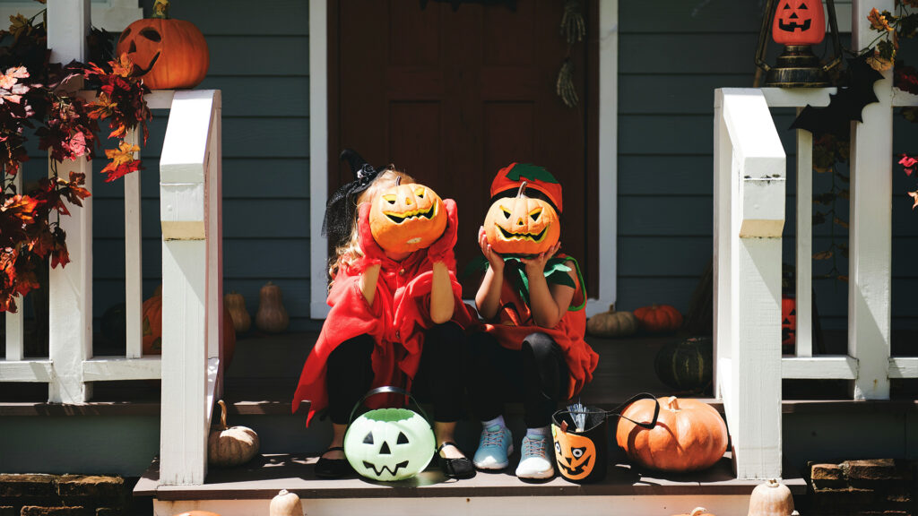 Two kids in adorable Halloween costumes holding pumpkin baskets, sitting on a porch surrounded by Halloween decorations.