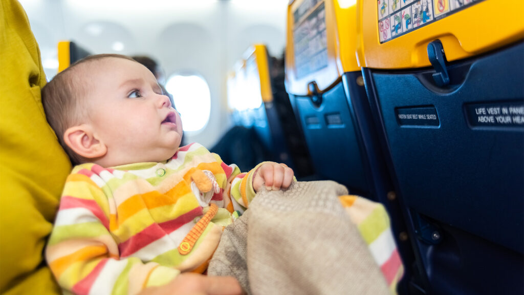 A baby in colorful clothes sitting on a parent's lap, gazing out of the window during a flight. Tips for flying with a newborn.