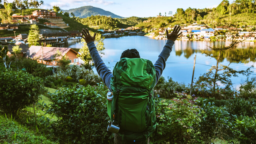 Traveler with a green backpack enjoying a scenic view of a lake and mountains – Sustainable Travel Tips for mindful and eco-friendly adventures.
