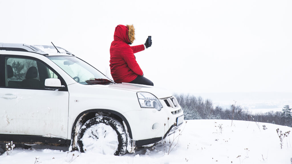 A person sitting on a white SUV, taking a selfie in a snowy winter landscape, with snow covering the ground and trees.