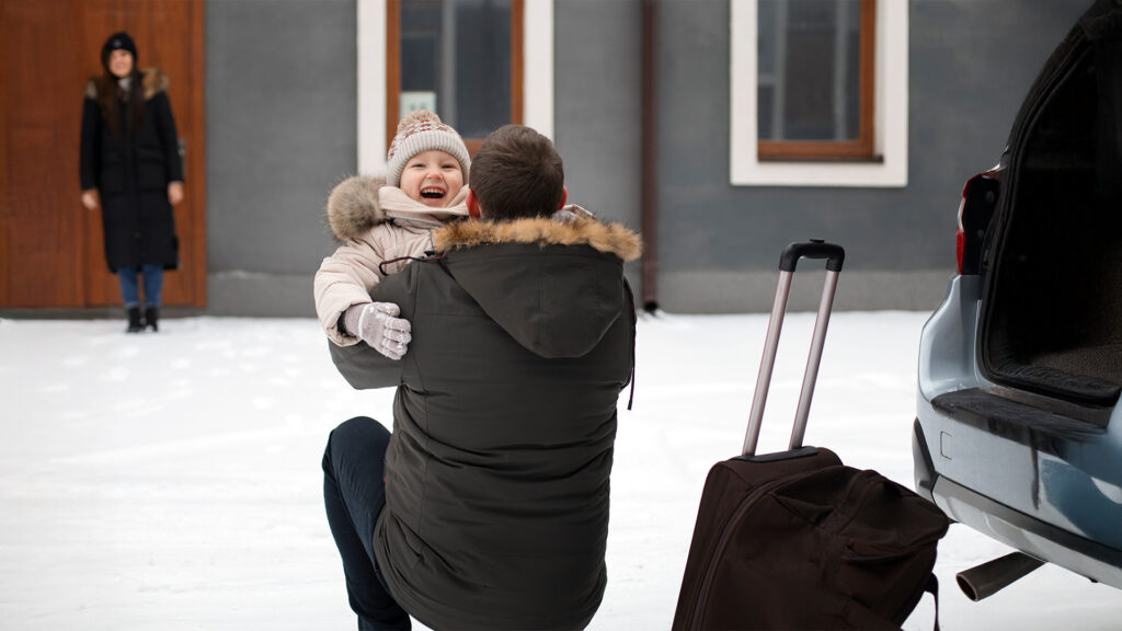 A father lifting his child in the snow, with luggage by their side, preparing for a winter vacation.
