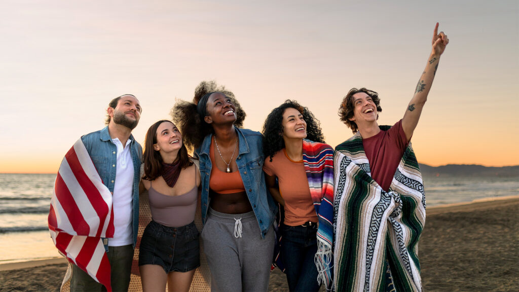 Diverse group of people smiling and standing together on a beach, wrapped in blankets with the American flag, celebrating unity and hope on MLK Day 2026 at the beach.