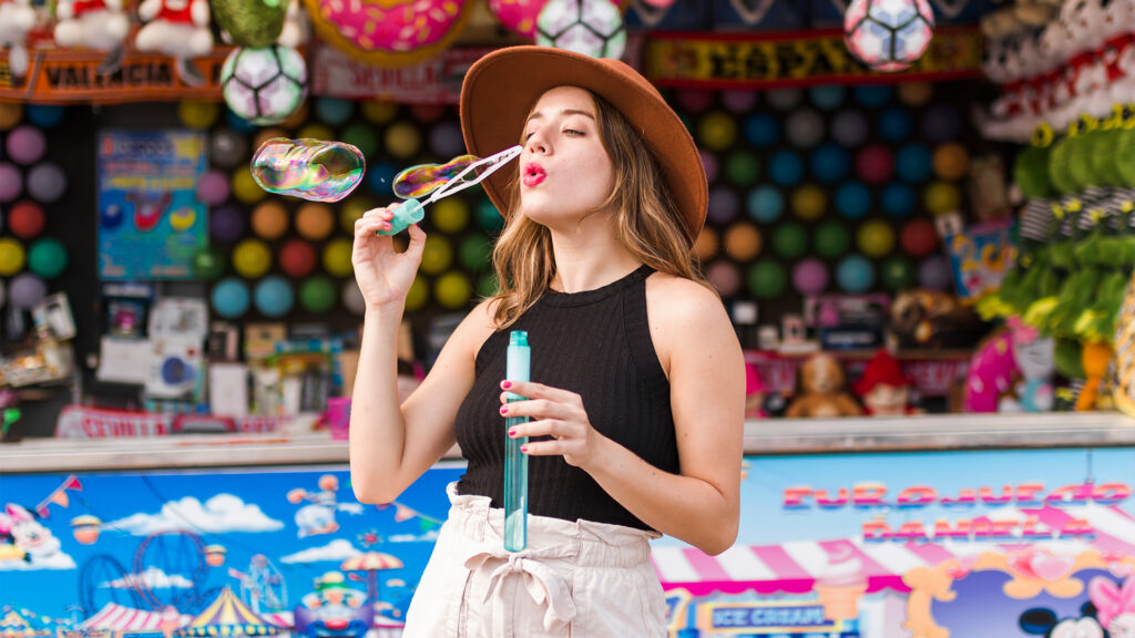 A woman enjoying a fun moment at an amusement park during February travel, blowing bubbles in front of vibrant stalls.