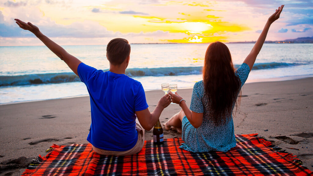 A couple raising glasses of champagne, watching the sunset on a beach with a scenic view of the ocean, perfect for a romantic getaway.
