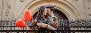 A couple holding hands and smiling, surrounded by romantic atmosphere with red heart-shaped balloons against a beautiful historic building.