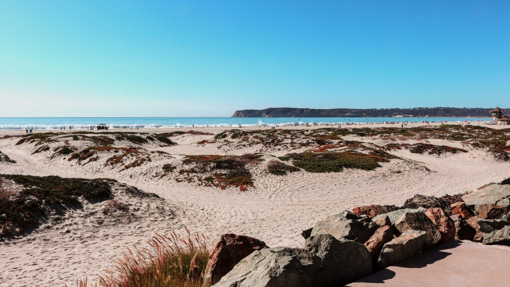 A wide beach with sandy dunes leading to the ocean, with clear skies and a view of the coastline stretching into the distance. Some people are enjoying the beach.