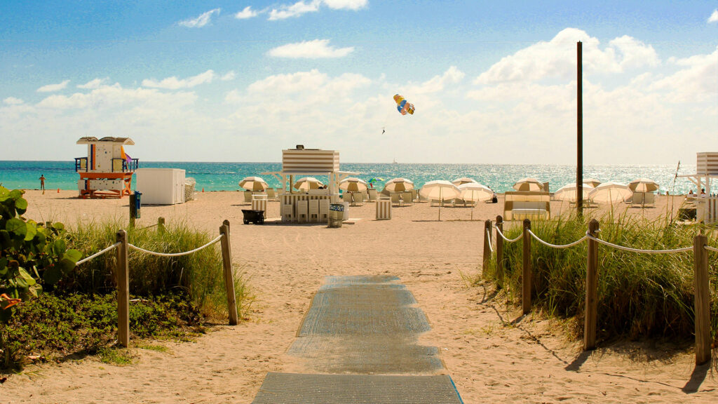 A peaceful beach path leading toward the water, with beach umbrellas and a lifeguard tower in the distance under a bright sunny sky.