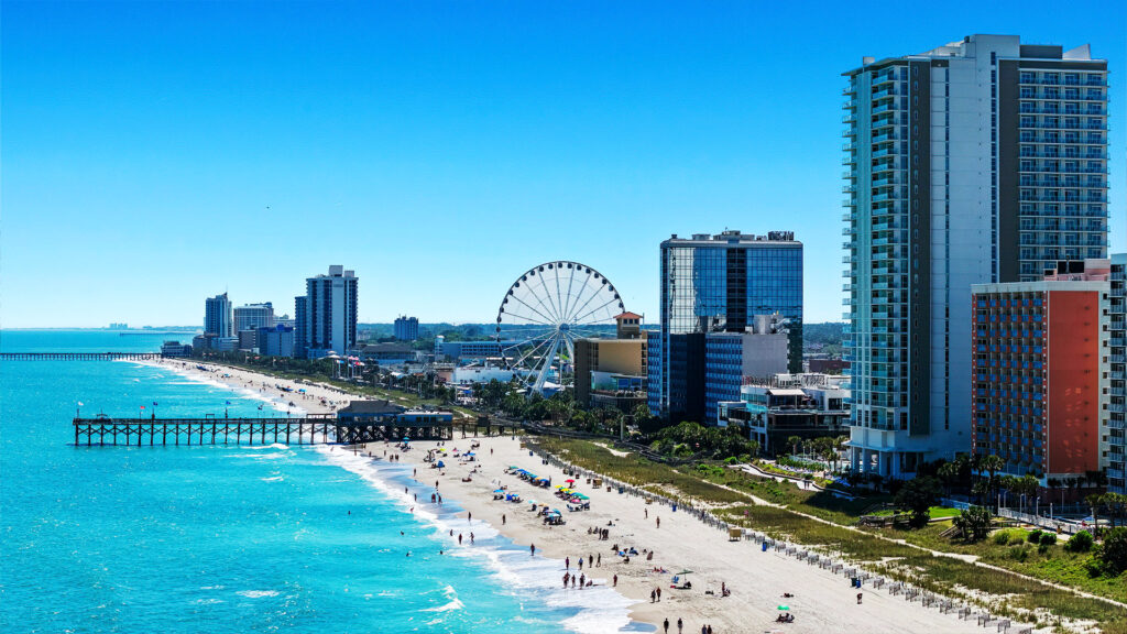 A lively beach town with high-rise buildings, a Ferris wheel, and people walking along the sandy shore under clear skies.