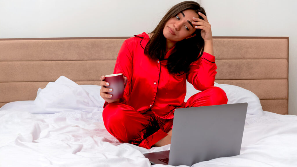 Woman feeling stressed while working on a laptop in bed, possibly dealing with last-minute hotel bookings for summer travel 2026.