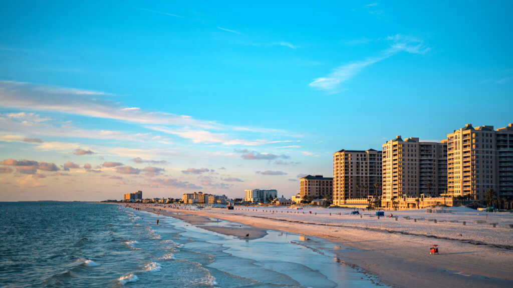 A beach town at twilight with soft golden hues in the sky, tall buildings lining the coastline, and the gentle waves rolling onto the shore.