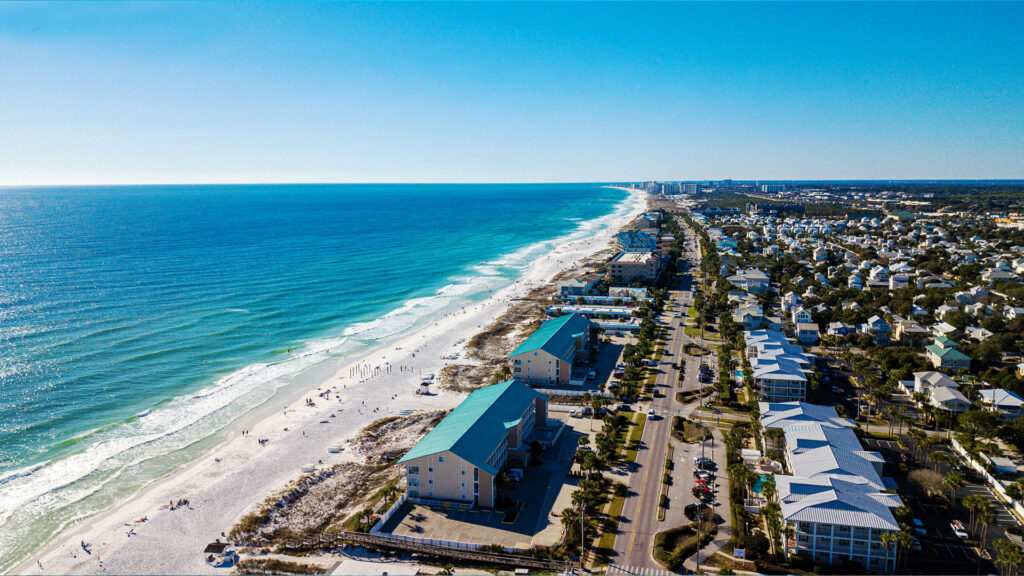 A beachside view with a stretch of sand, lush greenery, and clear waters, with buildings along the coastline and calm waves.