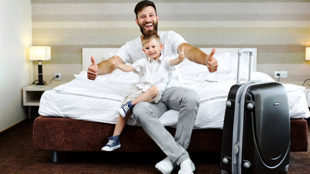 A father and son excited about their summer vacation, with their luggage packed for last-minute hotel booking in 2026.
