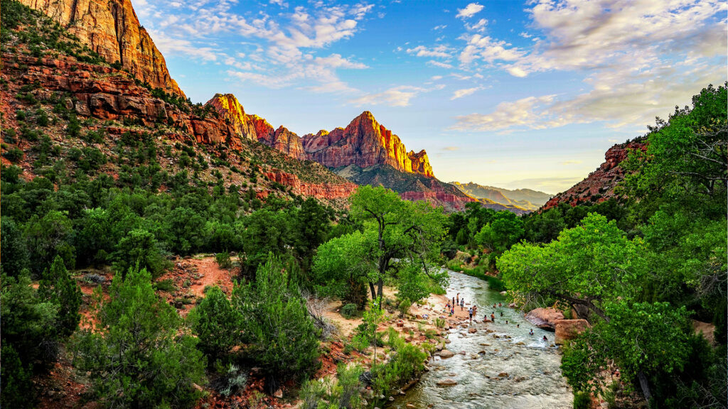 The dramatic red rock formations of Zion National Park, bathed in golden light with lush vegetation along the river.