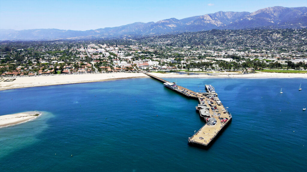 An aerial view of a pier extending into the ocean, with a town in the background and mountains in the distance. The crystal-clear water contrasts with the sandy beach.