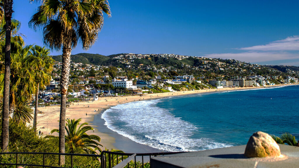 A beach lined with palm trees, overlooking the shoreline and hills beyond. The beach is calm with a few visitors enjoying the sun.