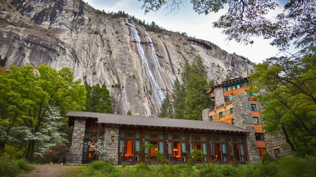 Historic stone lodge beneath a dramatic waterfall in Yosemite Valley showcasing iconic hotels near U.S. National Parks.