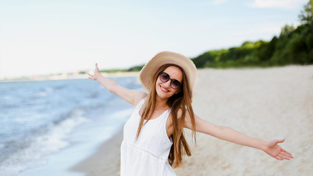 A smiling woman enjoying the beach breeze on a sunny day, capturing the essence of a laid-back summer vacation at an underrated destination.